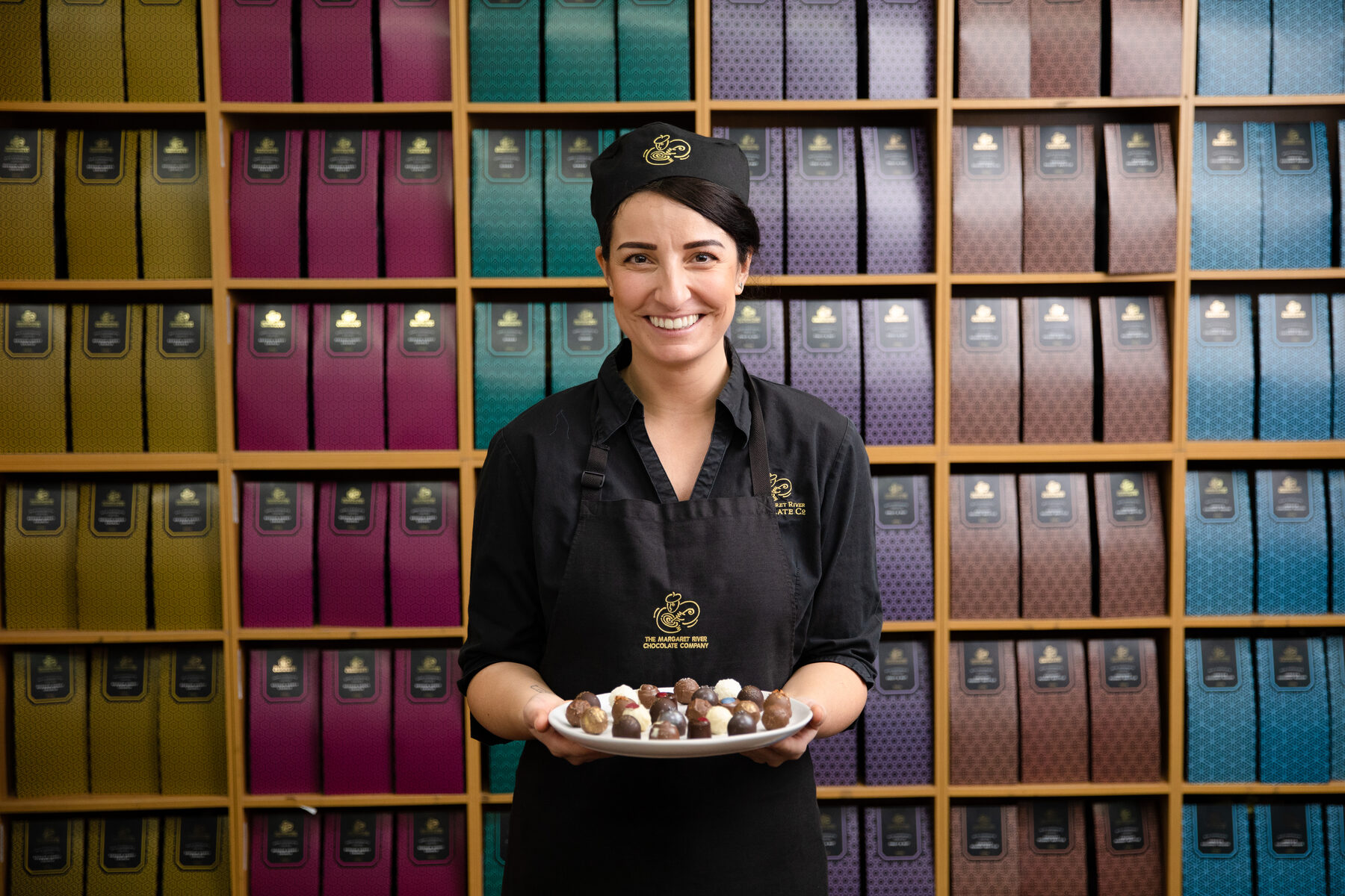 A chocolatier holding a tray of truffles in front of a wall of colourful chocolate blocks
